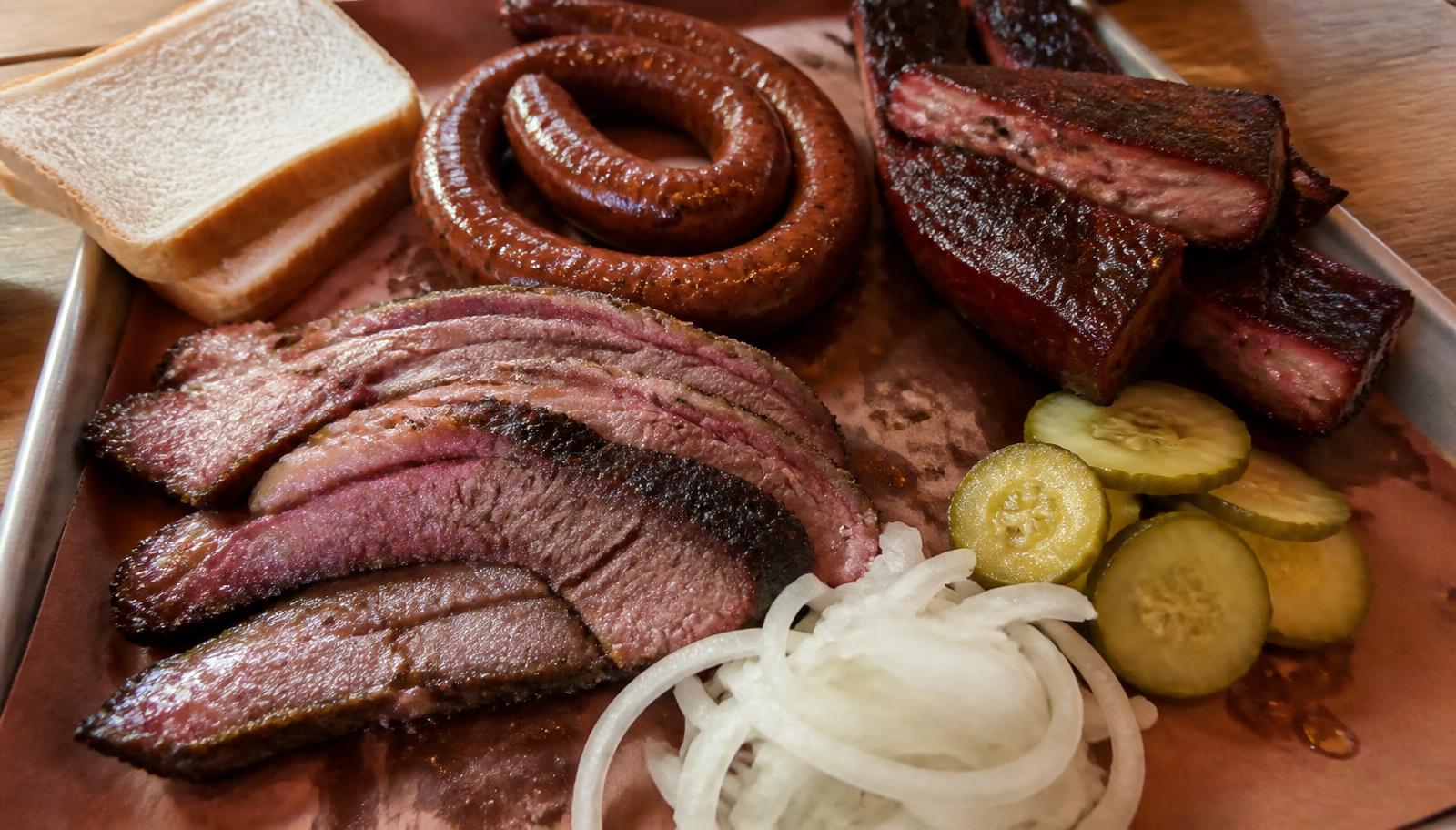 A Texas barbecue tray with sliced brisket, sausage, pork ribs, pickles, onions and white bread